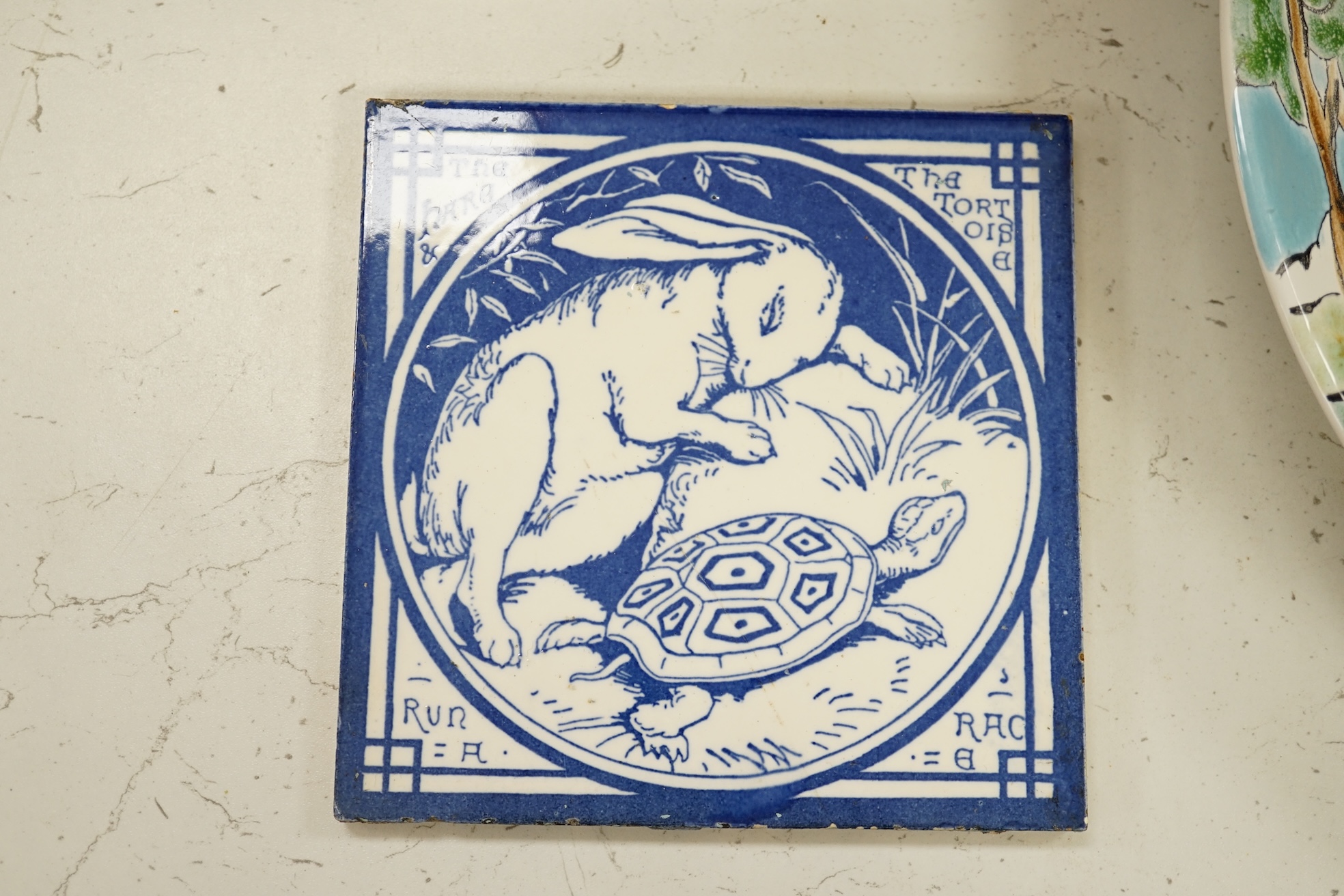 Two Minton pottery Arts and Crafts fireplace tiles, printed in blue and white, with scenes from Aesops fables - ‘’The Fox and the Goat in the Well’’ and ‘’The Hare and the Tortoise’’, together with a Poole pottery dish.